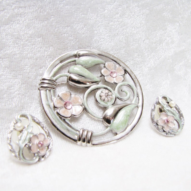 May include: A silver and green enamel brooch with pink flowers and rhinestones. Two matching earrings with the same design are also included.
