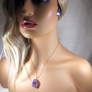 May include: A gold chain necklace with a purple amethyst crystal pendant. The pendant is a raw, natural crystal with a rough, irregular shape. The necklace is worn by a model with blonde hair.