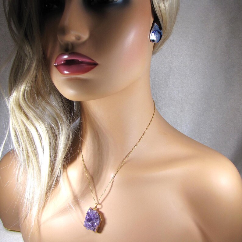 May include: A gold chain necklace with a purple amethyst crystal pendant. The pendant is a raw, natural crystal with a rough, irregular shape. The necklace is worn by a model with blonde hair.