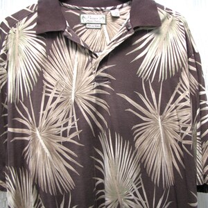 May include: Brown polo shirt with a tropical palm leaf pattern in shades of tan and beige. The shirt has a dark brown collar and a three-button placket. The tag reads "Paradise".