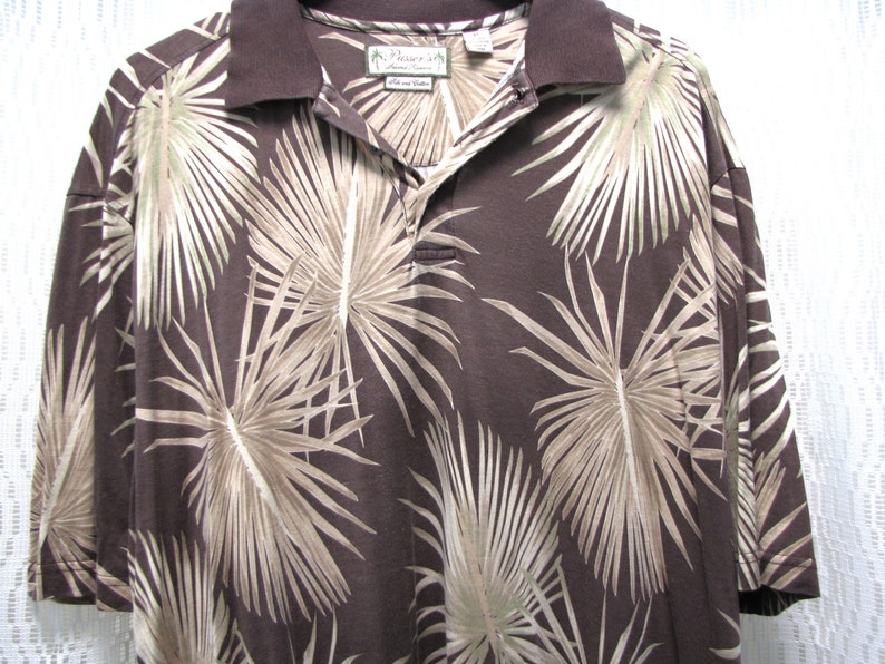 May include: Brown polo shirt with a tropical palm leaf pattern in shades of tan and beige. The shirt has a dark brown collar and a three-button placket. The tag reads "Paradise".