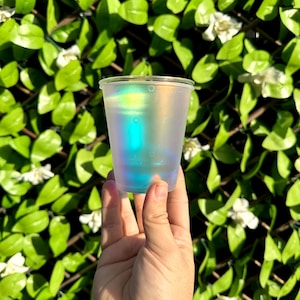 May include: A clear plastic cup with an iridescent finish. The cup is filled with a blue liquid and is being held by a hand.
