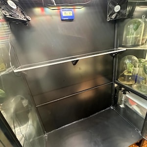 May include: An indoor plant growing cabinet with a black interior, featuring a clear shelf, two fans, and a digital temperature and humidity display. The cabinet has a built-in LED grow light and glass cloches on the right side.