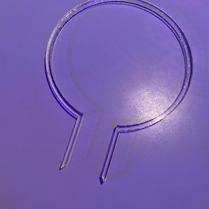 May include: A clear plastic ring with two prongs extending from the bottom. The ring is on a purple surface.
