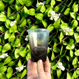 May include: A clear plastic cup with holes in the bottom, filled with dark brown soil. The cup is being held in a hand against a background of green leaves and white flowers.