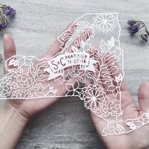 May include: White paper cut-out of the state of New York with floral designs and the text "S+C Married 10-27-18" in a banner.