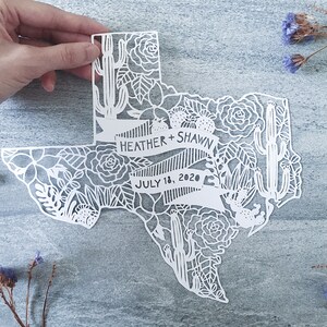 May include: White paper cut-out of the state of Texas with floral and cactus designs. The text "Heather + Shawn" and "July 18, 2020" is included in the design.