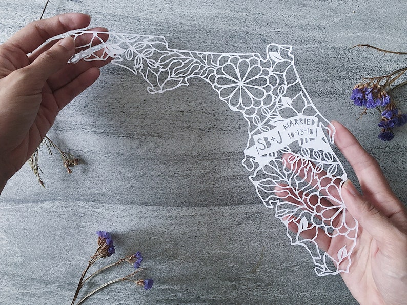 May include: White paper cut-out of the state of Florida with intricate floral designs. The text "SB+J Married 10-13-18" is on a banner in the lower right corner.