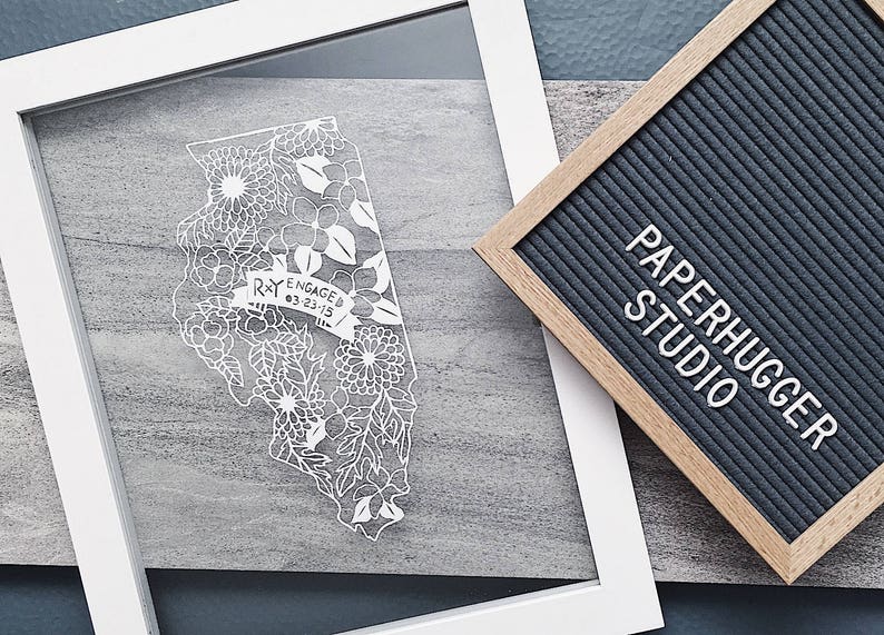 May include: White paper cut-out art of the state of Illinois with floral designs and the text "R+Y ENGAGED 03.23.15" in a banner. The art is framed in a white frame and displayed on a gray surface.