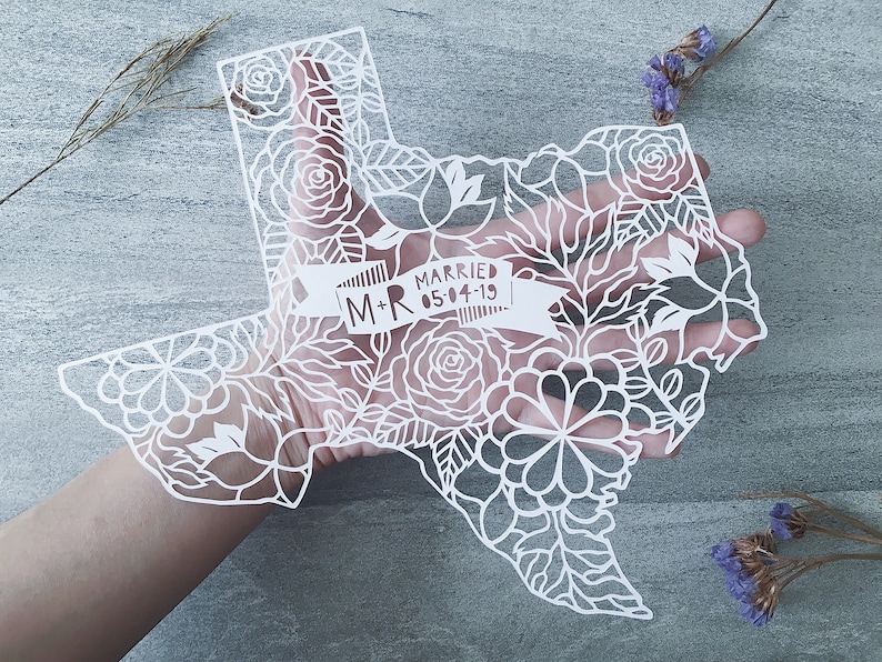 May include: White paper cut-out of the state of Texas with intricate floral designs and a banner that reads "M+R Married 05-04-19".