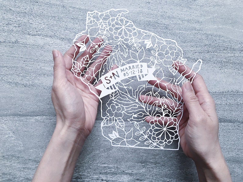 May include: White paper cut-out of the state of Wisconsin with floral designs and the text "S+N Married 05-12-18" on a banner.