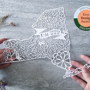 May include: White paper cut-out of the state of New York with intricate floral designs. The design includes a banner with the text "R+M Married 01-18-15".