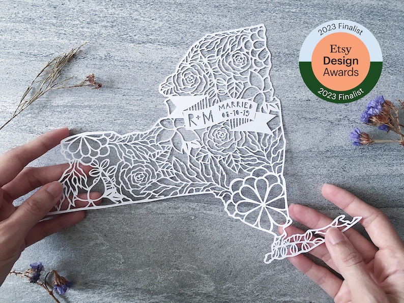 May include: White paper cut-out of the state of New York with intricate floral designs. The design includes a banner with the text "R+M Married 01-18-15".