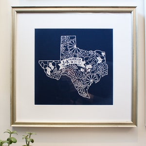 May include: A framed print of a white paper cut-out of the state of Texas with floral details. The print is on a dark blue background and includes the text "J+K Married 10-21-17".