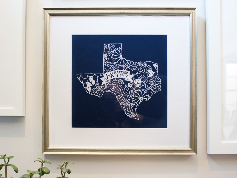 May include: A framed print of a white paper cut-out of the state of Texas with floral details. The print is on a dark blue background and includes the text "J+K Married 10-21-17".