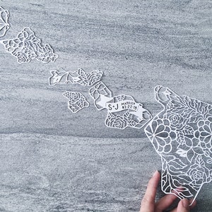 May include: White paper cut-out of the Hawaiian islands with floral designs. The islands are arranged in a map shape with a banner that reads "S+J Married 03.22.19".