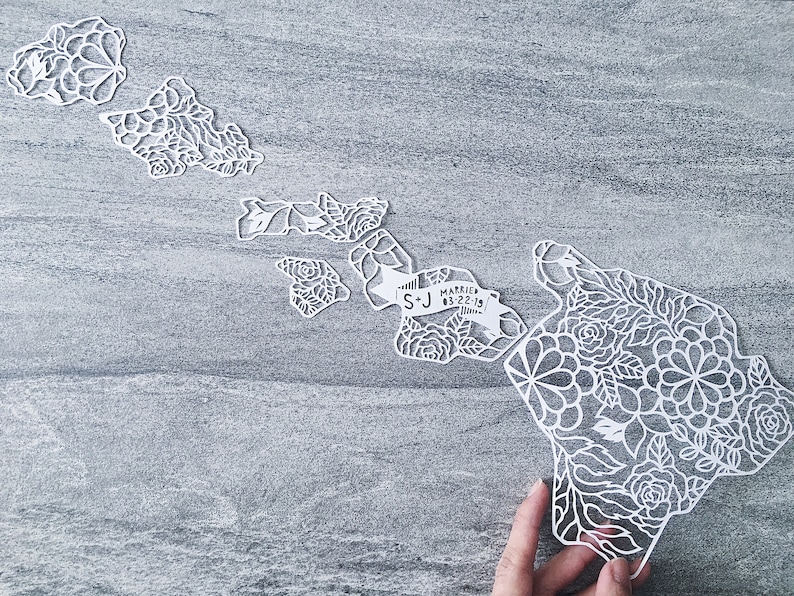 May include: White paper cut-out of the Hawaiian islands with floral designs. The islands are arranged in a map shape with a banner that reads "S+J Married 03.22.19".