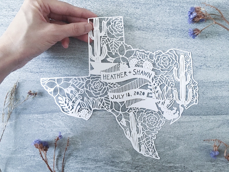 May include: White paper cut-out of the state of Texas with floral and cactus designs. The text "Heather + Shawn" and "July 18, 2020" is included in the design.