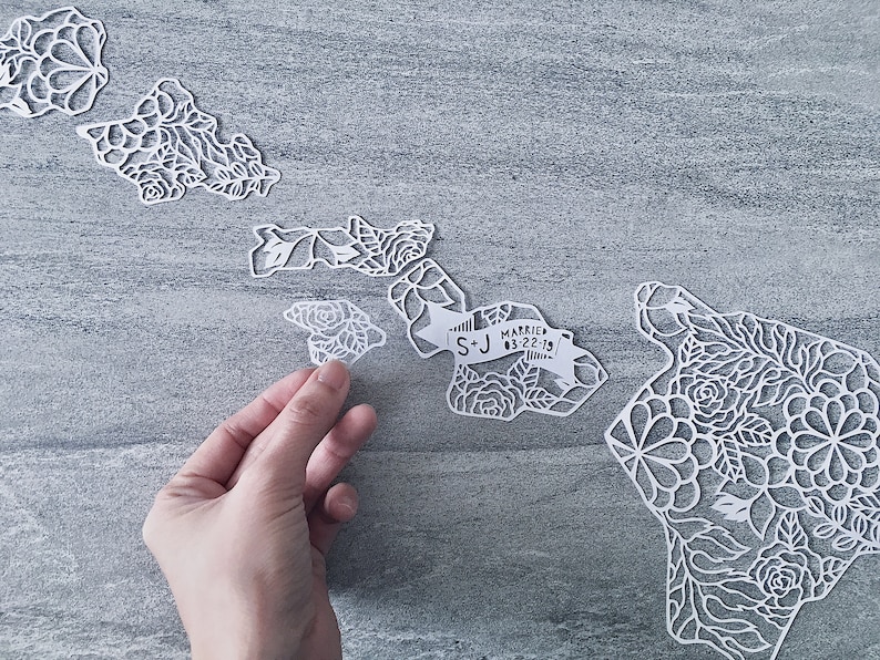 May include: White paper cut-out of the Hawaiian islands with intricate floral designs. The islands are arranged in a line, with a banner reading "S+J Married 03-22-19" on the largest island.