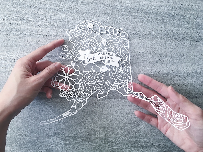 May include: White paper cut-out of the state of Alaska with floral designs and the text "S+E Married 01-01-18" in a banner.