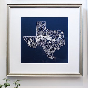 May include: A white paper cut-out of the state of Texas with floral designs and the text "J+K Married 10.21.17" on a blue background. The paper cut-out is framed in a silver frame.