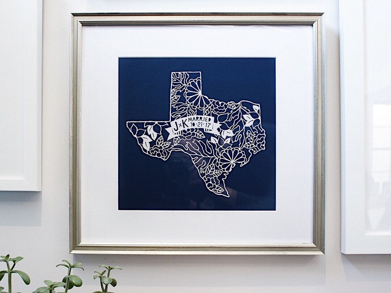 May include: A white paper cut-out of the state of Texas with floral designs and the text "J+K Married 10.21.17" on a blue background. The paper cut-out is framed in a silver frame.