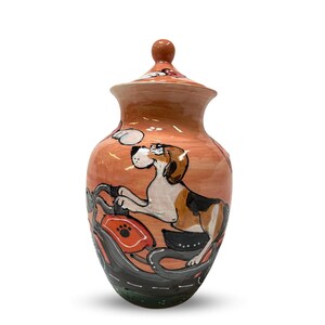 May include: A ceramic urn with a peach-colored base and a lid. The urn is decorated with a hand-painted illustration of a beagle dog riding a motorcycle. The dog is wearing a black helmet and goggles. The motorcycle is red and black.