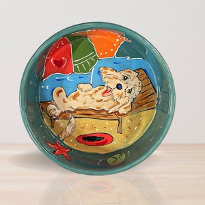 May include: A teal ceramic dog bowl with a hand-painted design of a dog relaxing on a beach chair under an umbrella. The dog is light brown with a blue nose and tongue sticking out. The bowl has a starfish, a red and yellow beach ball, and a green ball on the side.