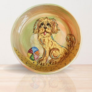 May include: Hand-painted ceramic dog bowl featuring a golden-colored dog with a colorful ball. The bowl has a light green, orange, and yellow background. The dog is sitting with its tongue out. The bowl is round and has a glossy finish.