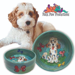 May include: Two teal blue ceramic dog bowls with a hand-painted design of a dog with white and brown fur. The larger bowl has a yellow bow and red bone designs. The smaller bowl has red bone designs. The bowls are on a white background with a dog in the background.