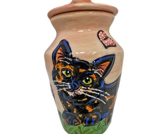 May include: A ceramic pet urn with a hand-painted design of a black and orange cat with green eyes. The urn has a pink lid with a white heart design. The cat is sitting on a green grass background.