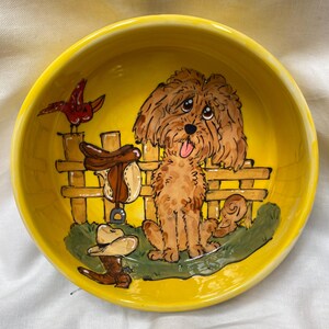 May include: A yellow ceramic dog bowl with a hand-painted design of a brown dog sitting in front of a wooden fence with a saddle and a red bird on top. The dog has a brown and white coat and is looking at the viewer.