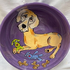 May include: Purple ceramic dog bowl with a hand-painted image of a yellow dog with brown spots, a green toy, and a blue toy. The bowl also has brown bone designs around the rim.