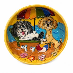 May include: A yellow ceramic bowl with a beach scene featuring two cartoon dogs. One dog is lounging on a beach chair under a rainbow umbrella, while the other stands beside a beach ball. The bowl is decorated with seashells and starfish.