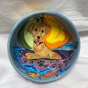 May include: A hand-painted ceramic bowl with a light blue rim and a whimsical design. The bowl features a golden retriever in a boat, with colorful waves, fish, and a yellow sail. The interior is painted with vibrant colors.
