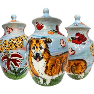 May include: Three ceramic urns with lids, each featuring a different painted design. The center urn has a brown and white dog with a red tongue, surrounded by flowers and hearts. The left urn has a leopard print design and the right urn has a red bird design.