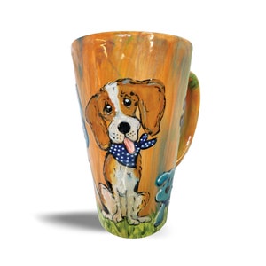 May include: A ceramic mug with a hand-painted design of a beagle dog wearing a blue bandana. The mug has a brown, orange, and yellow background with green grass and blue flowers.