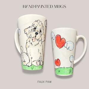 May include: Two hand-painted ceramic mugs. One mug features a white dog with brown markings sitting on a green background. The other mug has a white background with red hearts and white wings. Both mugs have the text "FAUX PAW" printed on the bottom.