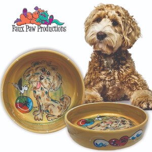 May include: A custom-painted ceramic dog bowl with a light brown background. The bowl features a cartoon illustration of a dog with a ball and a bird. The text "Faux Paw Productions" is visible in the top left corner of the image.