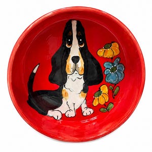 May include: A red ceramic bowl featuring a hand-painted illustration of a Basset Hound dog. The dog is black, white, and tan, with large, droopy ears and sad eyes. Yellow and blue flowers are painted on the bowl's interior.