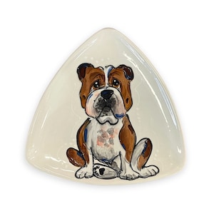 May include: A white triangular ceramic dish with a hand-painted illustration of a brown and white bulldog sitting on its hind legs. The bulldog has a black nose and blue eyes.
