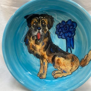 May include: A blue ceramic dog bowl with a hand-painted image of a brown and black dog with a blue ribbon. The dog is sitting and looking to the right.