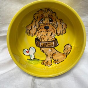 May include: A yellow ceramic dog bowl with a hand-painted illustration of a cartoon dog wearing a brown collar that says "Chewy Vuitton." A white bone with a red heart is next to the dog. The bowl is round.