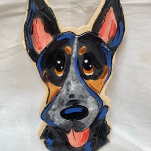 May include: Hand-painted ceramic tile featuring a cartoon dog with blue, black, and gray markings. The dog has a red tongue sticking out and large brown eyes.