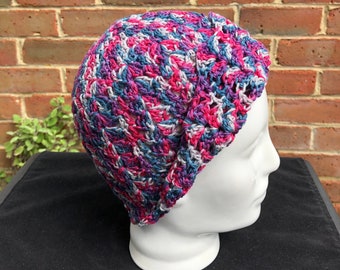 May include: A crocheted hat in a multi-colored pattern of pink, blue, and gray. The hat is on a white mannequin head.