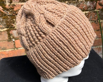 Puede incluir: Un gorro tejido de color beige. El gorro presenta un borde acanalado y un diseño de trenzas en la parte superior. Está hecho de un material suave y cálido, ideal para el frío. El gorro se muestra en una cabeza de maniquí blanca.