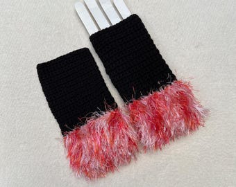 May include: A pair of black crochet wrist cuffs with a vibrant, fluffy fringe in shades of pink, red, and white. The cuffs are displayed on a white surface, showcasing the texture and color contrast of the handmade accessories.