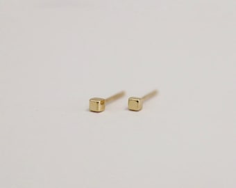 May include: A pair of small, square-shaped gold stud earrings. The earrings are simple and minimalist in design, with a polished finish. They are set against a plain white background.