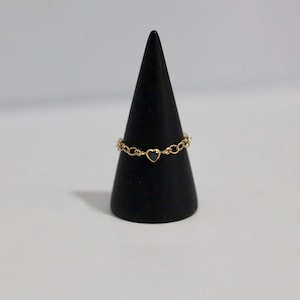 May include: A delicate gold ring with a heart-shaped center stone and a chain-link band. The ring is displayed on a black cone-shaped stand against a white background. The ring's design is simple and elegant.
