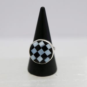 May include: A silver ring with a round face featuring a checkerboard pattern of black and light blue squares. The ring is displayed on a black cone-shaped stand against a white background. The ring's design is a bold, geometric statement.